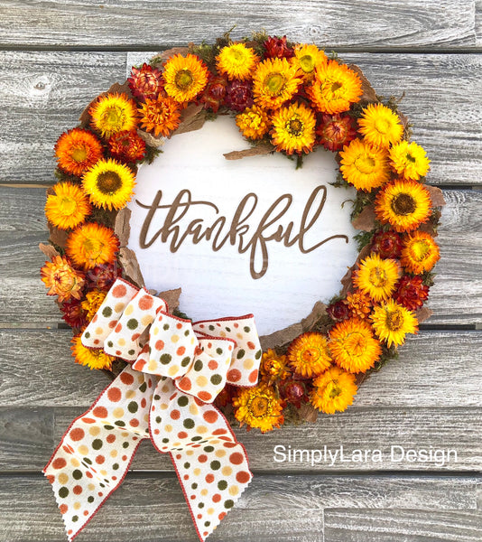 Autumnal wreath made of fall dried flowers, tree bark, green moss and a polka dot ribbon in coordination colors of orange, red, yellow, and green. Whitewashed wood background inscribed with the word Thankful. 